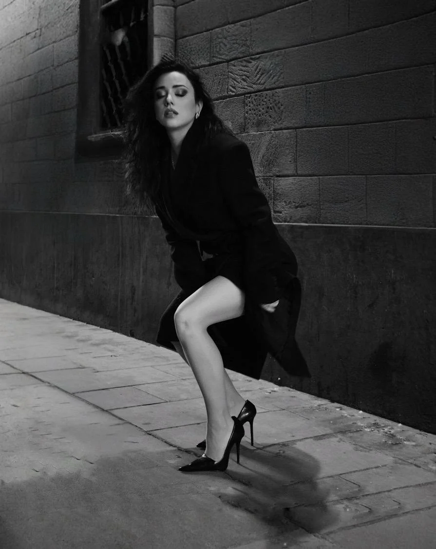 Mujer de cabello largo y oscuro en un callejón, usando un abrigo oscuro y tacones negros, en una pose dramática y elegante en una fotografía en blanco y negro.