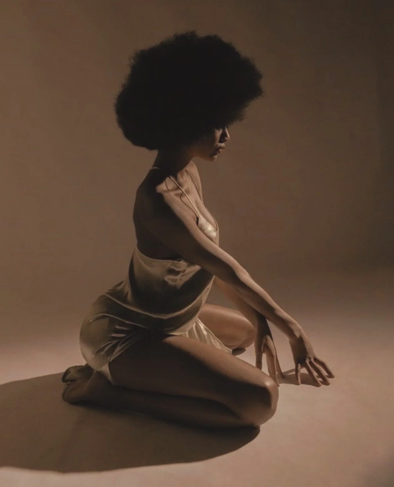 Mujer con cabello afro, en posición de rodillas, tocando el suelo con las manos en un fondo de color beige