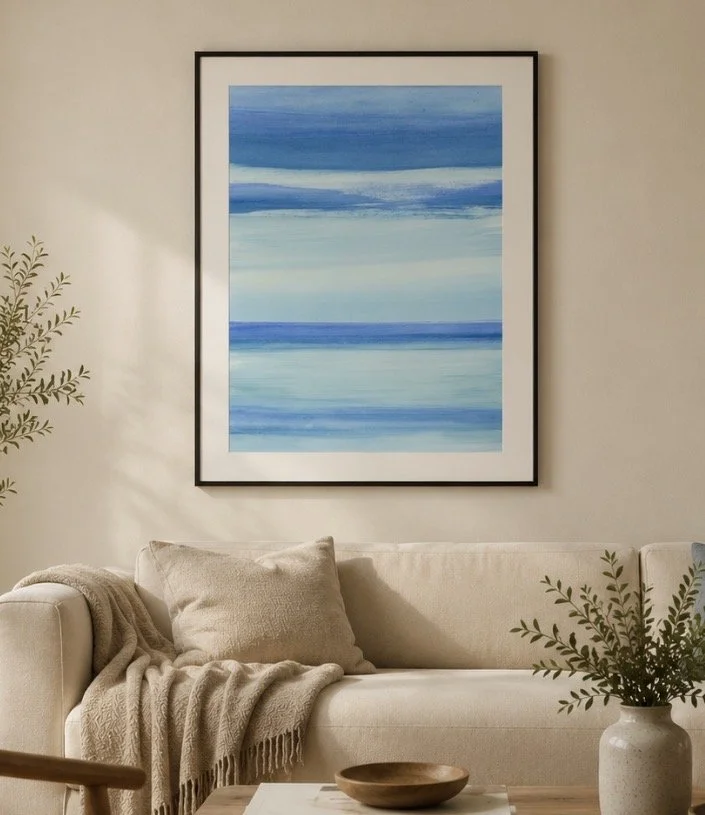 Cuadro abstracto con tonos azules en una pared del salón, con sofá beige, cojines, manta y plantas decorativas.