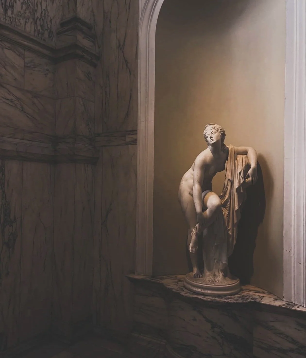 Escultura de una figura femenina en un pedestal, en un espacio con paredes de mármol y una pequeña pared de fondo de color beige.