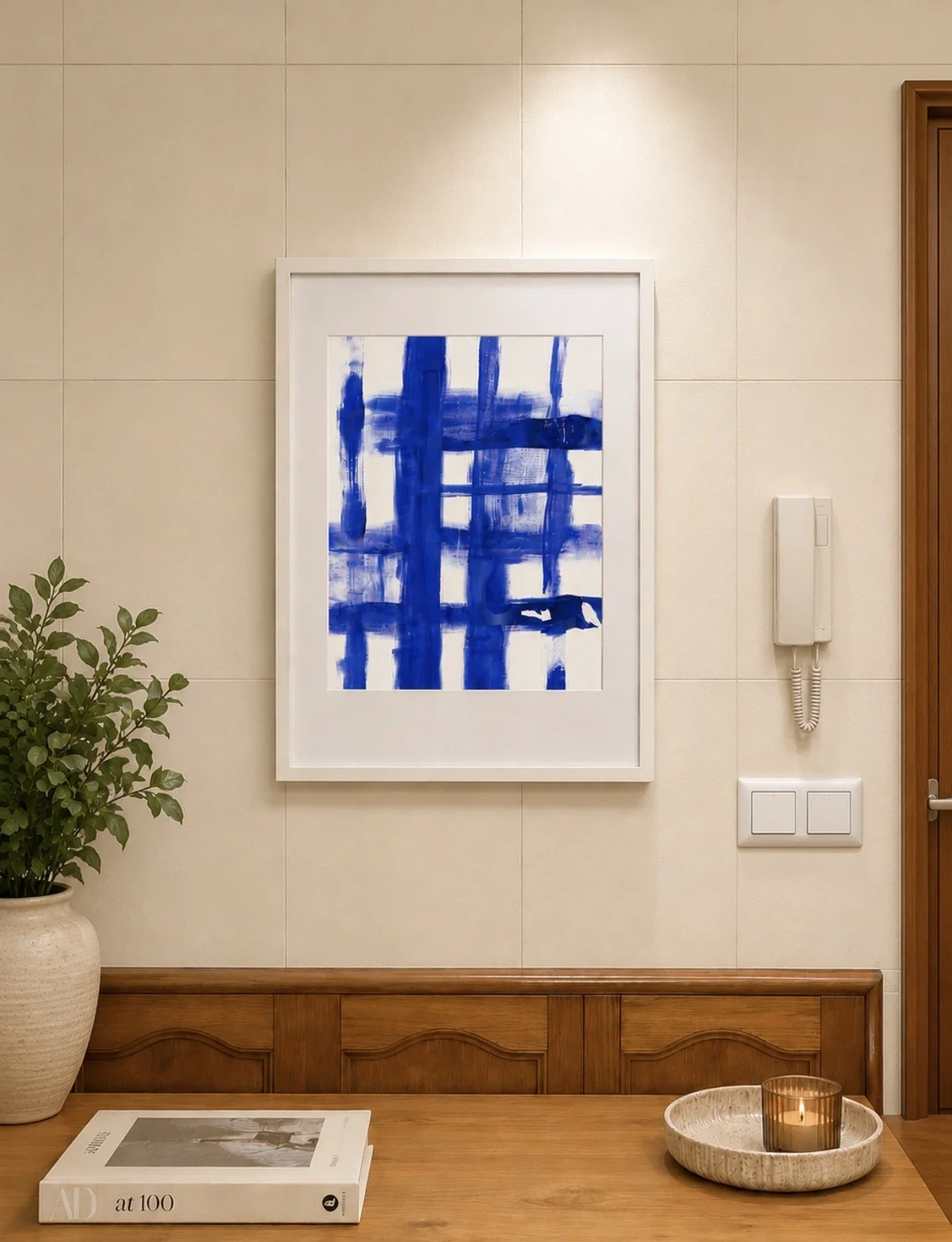 Pintura abstracta en tonos azules enmarcada en color blanco, colgada en pared crema. Planta en maceta a la izquierda, libro en mesa debajo de la pintura, y candelabro con vela en bandeja de cerámica al centro.