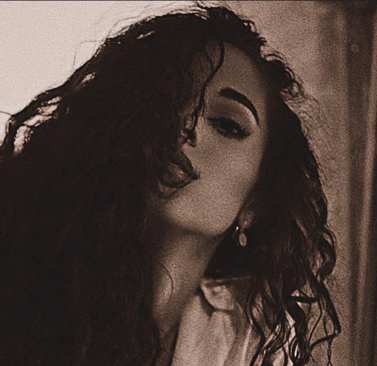 Retrato en sepia de una mujer con cabello rizado y maquillaje destacado, vistiendo un pendiente y una prenda blanca, con expresión pensativa