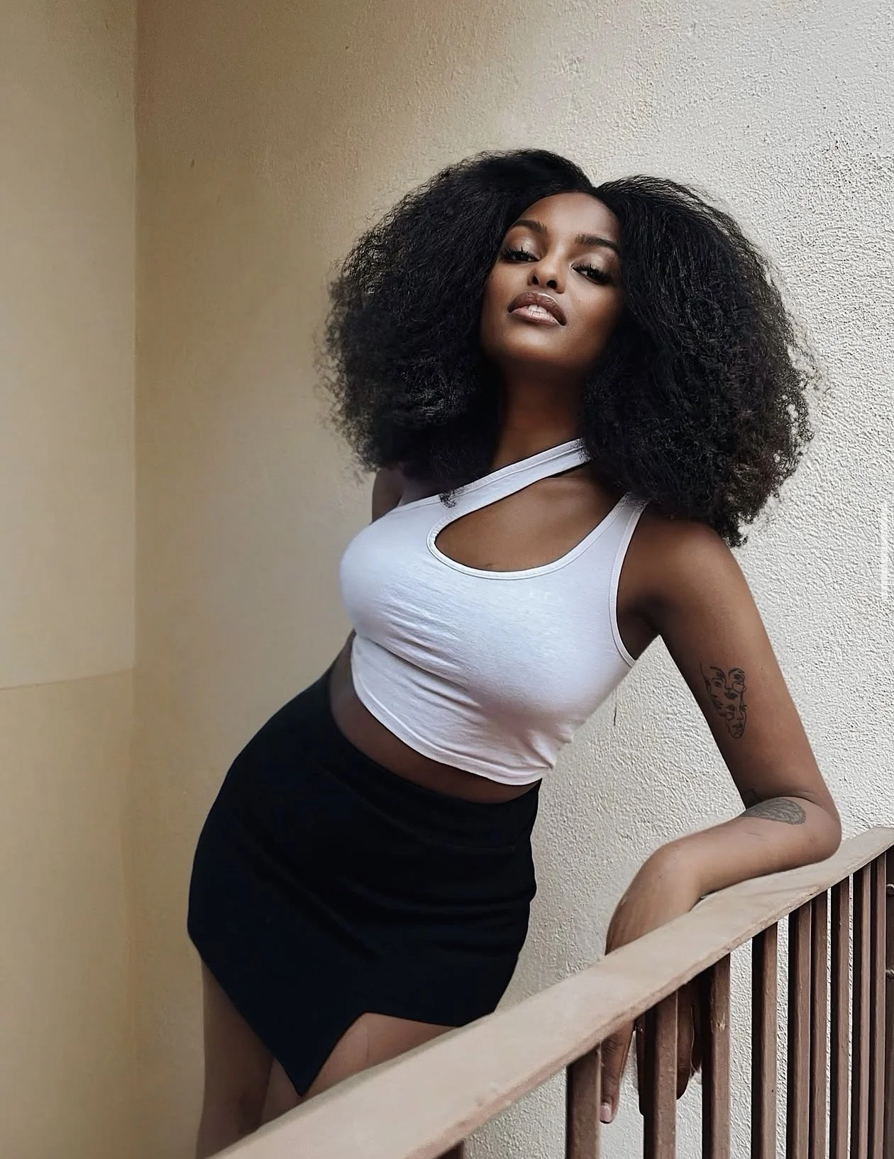 Una mujer con cabello rizado negro, usando una camiseta blanca sin mangas y una falda negra, apoyada en una barandilla de madera, posando frente a una pared de color claro.