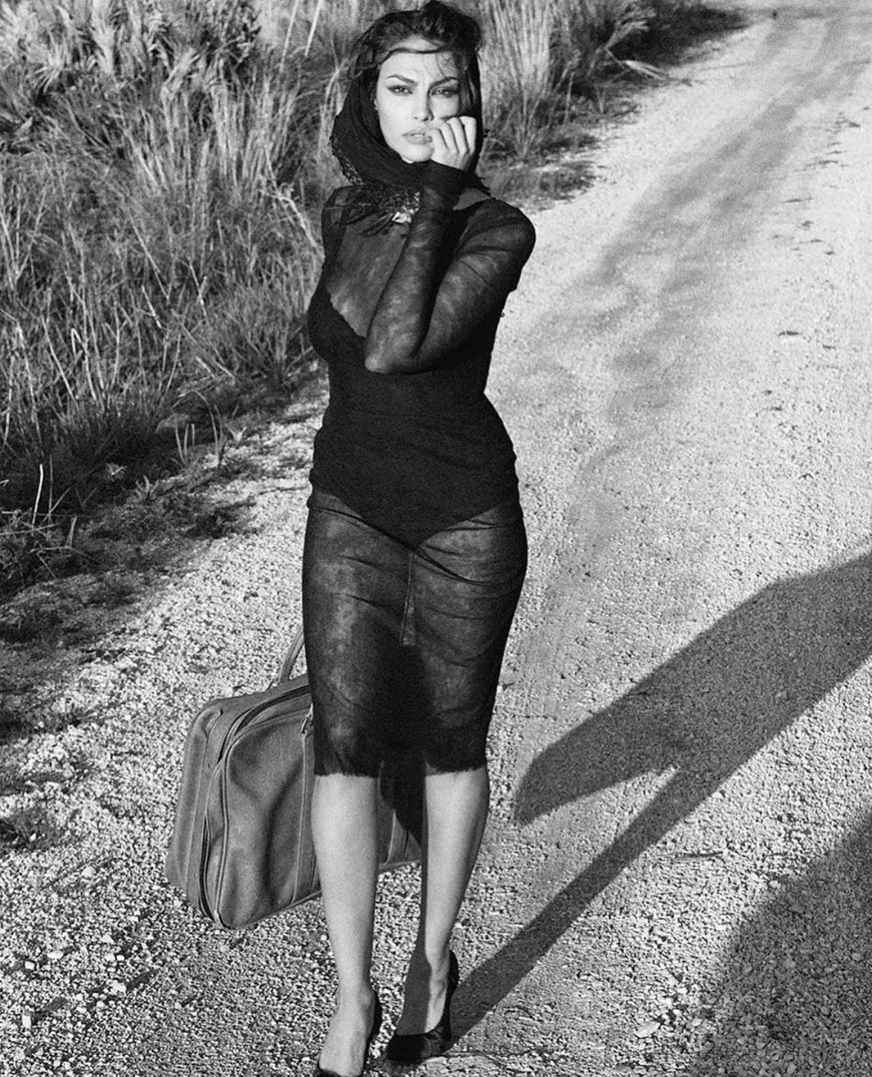 Mujer con vestido negro y bufanda, sosteniendo una maleta en una caminera de tierra, en un entorno al aire libre, en blanco y negro.