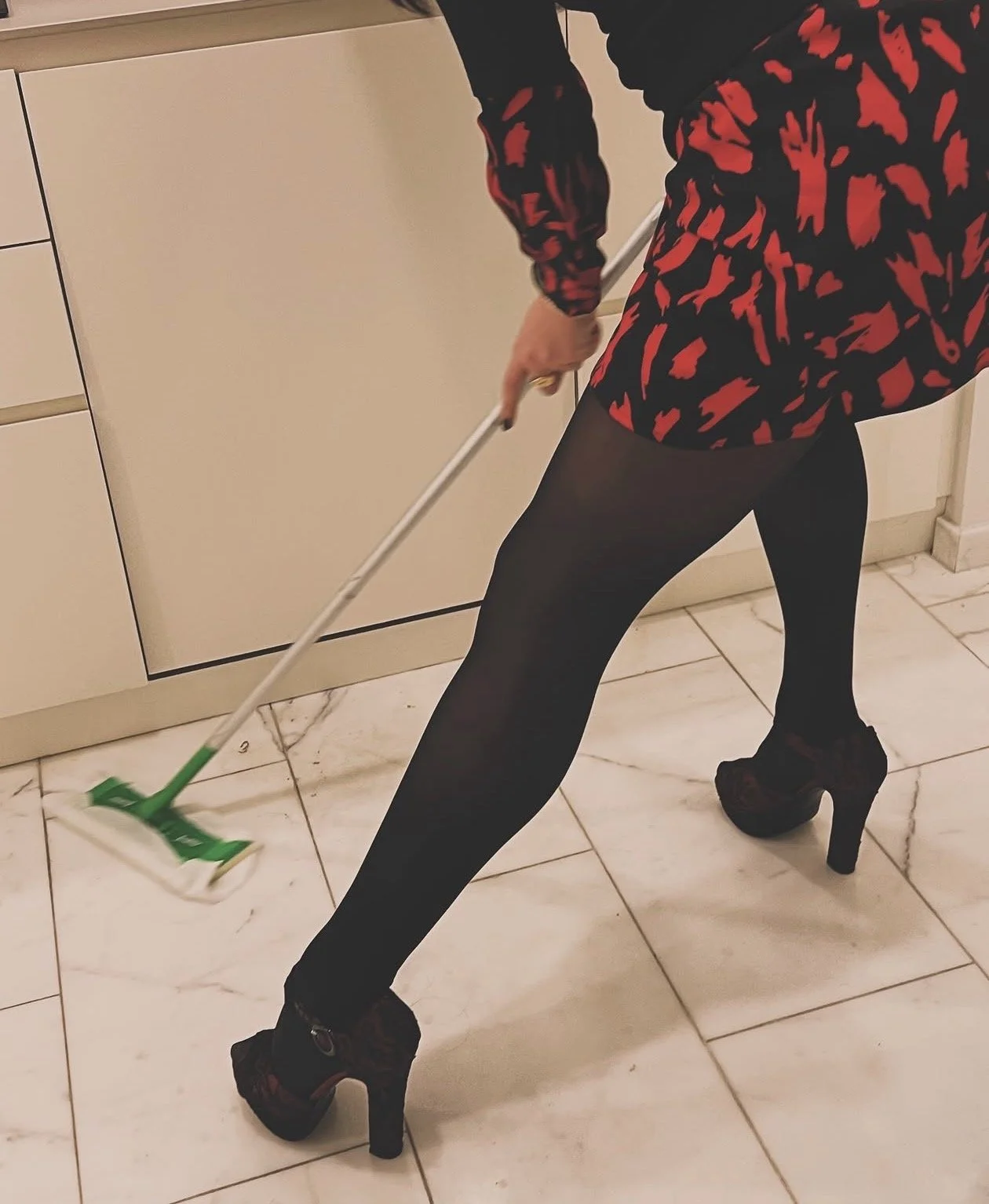 Persona con falda de animal print rojo y negro, calcetas negras, tacones altos y usando un trapeador en el suelo de un interior.