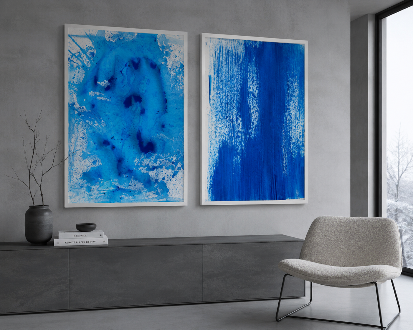 Dos cuadros abstractos azules en una pared de cemento en una sala moderna con un sillón beige y una mesa con libros y una vasija decorativa.