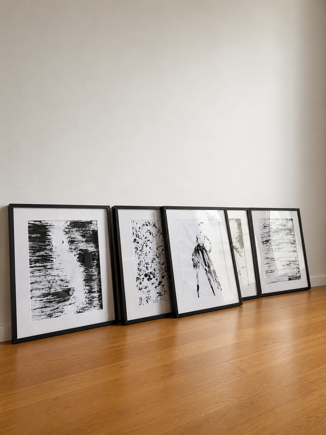 Cuatro cuadros negros con marcos apoyados en el suelo de una habitación con pared blanca y piso de madera, con arte en blanco y negro.