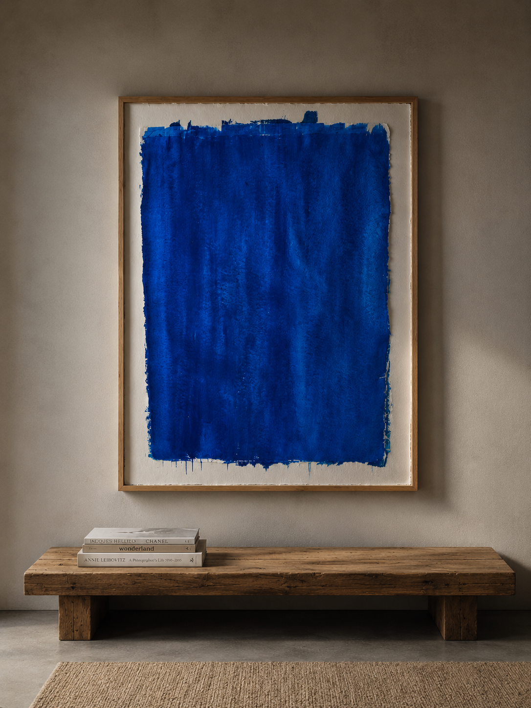 Cuadro abstracto de color azul en un marco de madera, colgado en una pared beige y descansando sobre un banco de madera, con libros apilados a la izquierda.