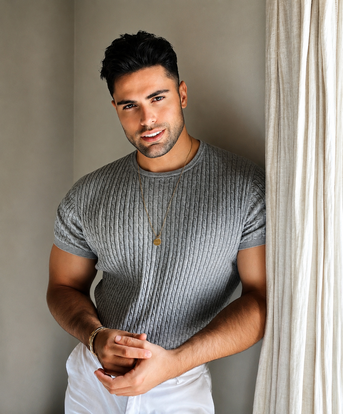 Joven hombre con cabello oscuro, barba, usando camiseta gris, collar y pulsera, posando junto a cortinas blancas en interior bien iluminado.