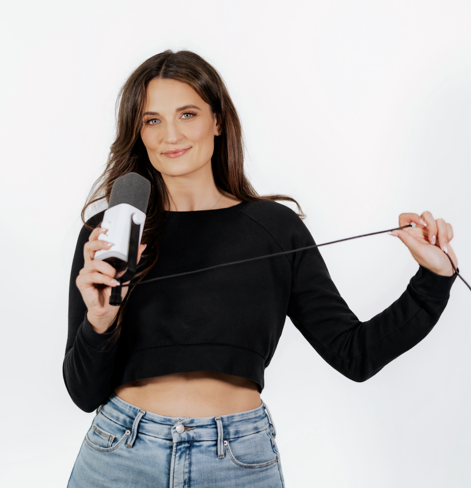 Woman with long brown hair smiling, holding a microphone in her right hand and pulling a cord with her left hand, wearing a black crop top and blue jeans against a white background.