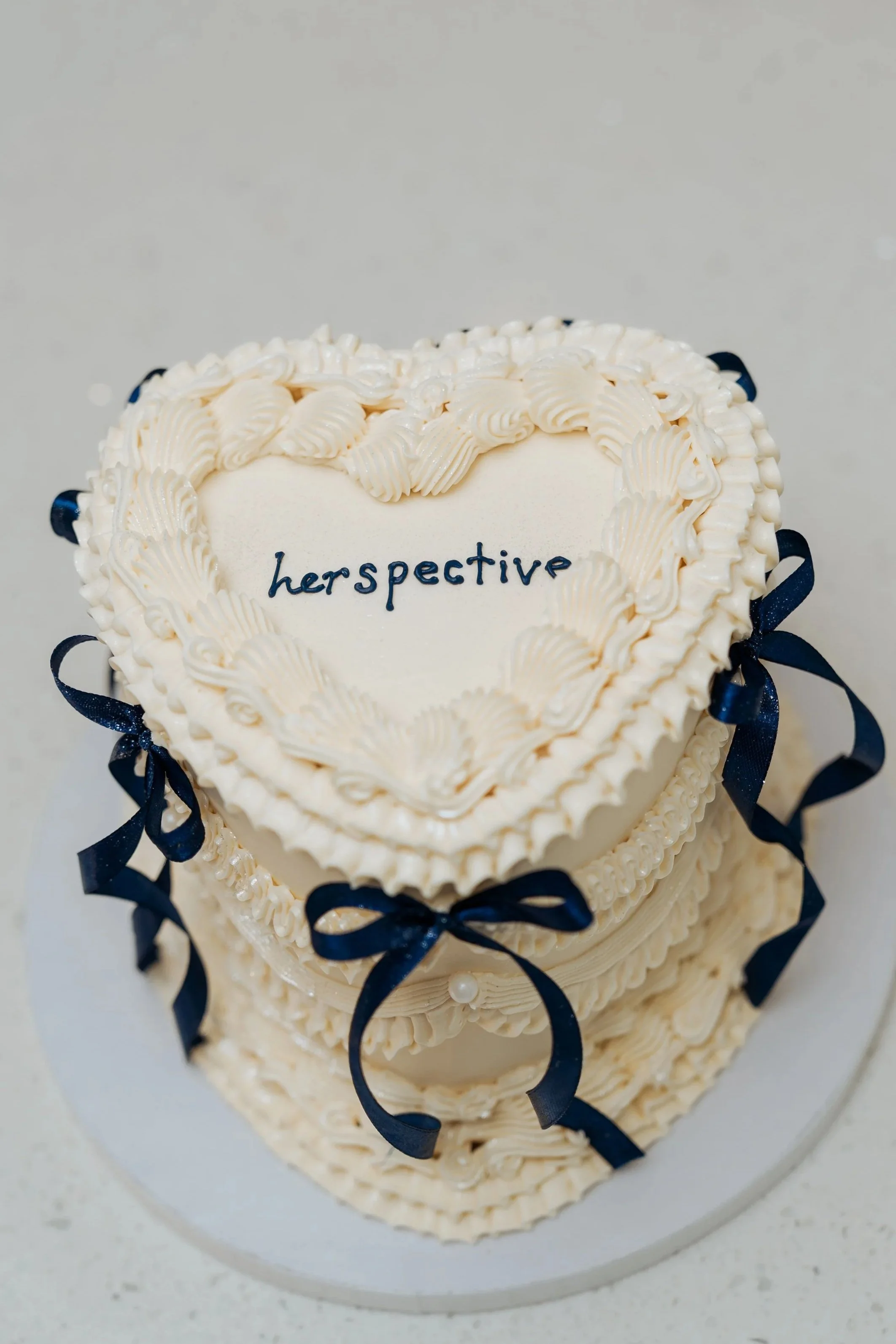 Close-up of a multi-tiered cake decorated with white icing and navy blue ribbons, with the word "perspective" written on the top tier in dark icing.