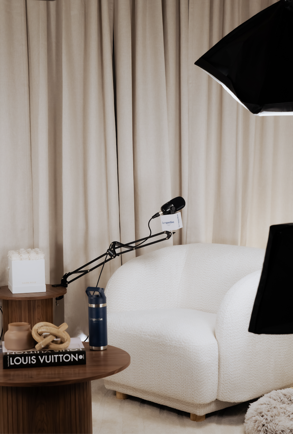 A cozy living room corner featuring a cream-colored textured sofa, a round wooden side table with decorative items including a Louis Vuitton book, a beige knot sculpture, a tan vase, and a blue water bottle. There's also professional lighting equipment directed at the sofa, with beige curtains in the background.
