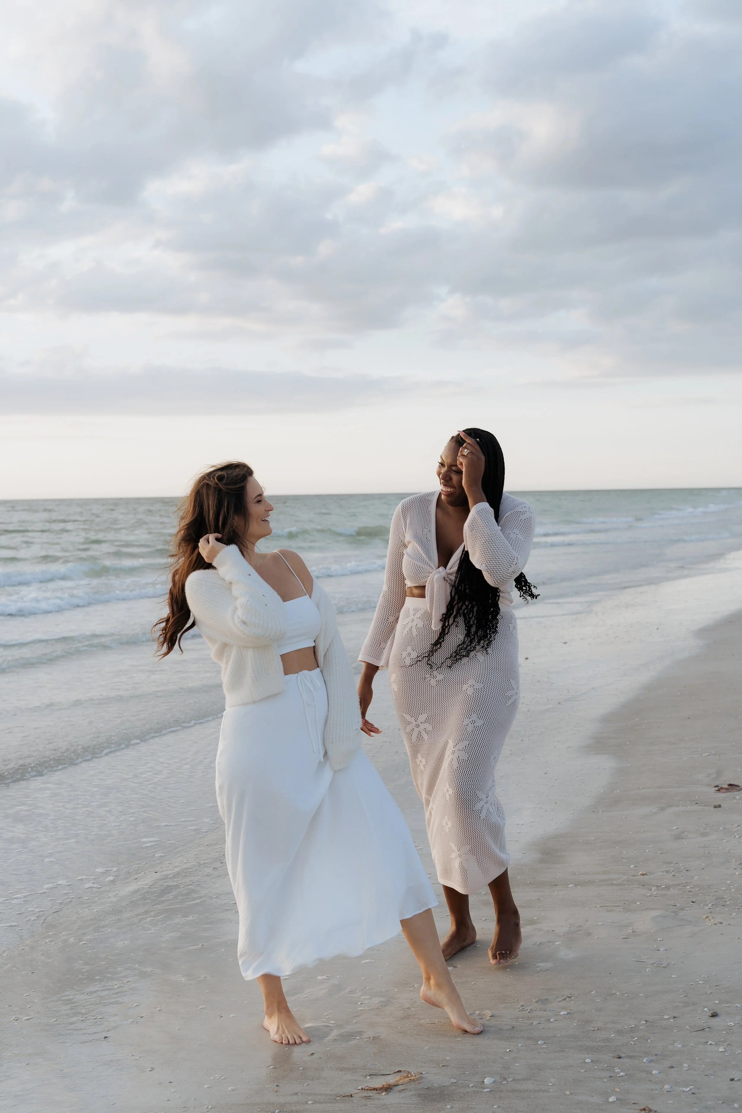 Two women enjoying a walk on the beach, dressed in white and beige outfits, smiling and laughing under cloudy skies.