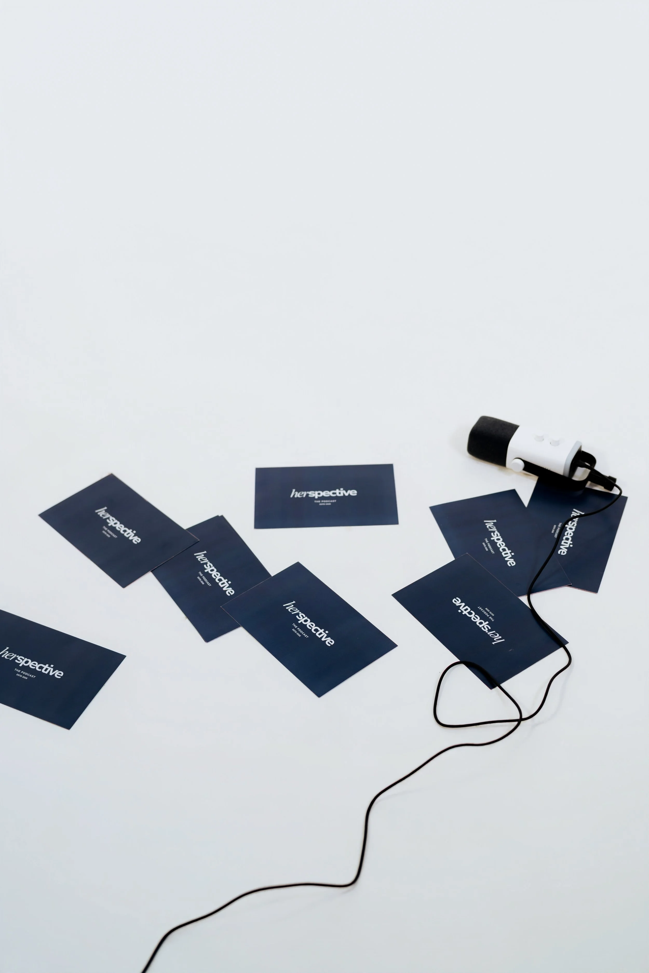 Scattered navy blue cards with white text that reads 'her perspective' on a white surface, with a black and white portable speaker connected to a black charging cable.