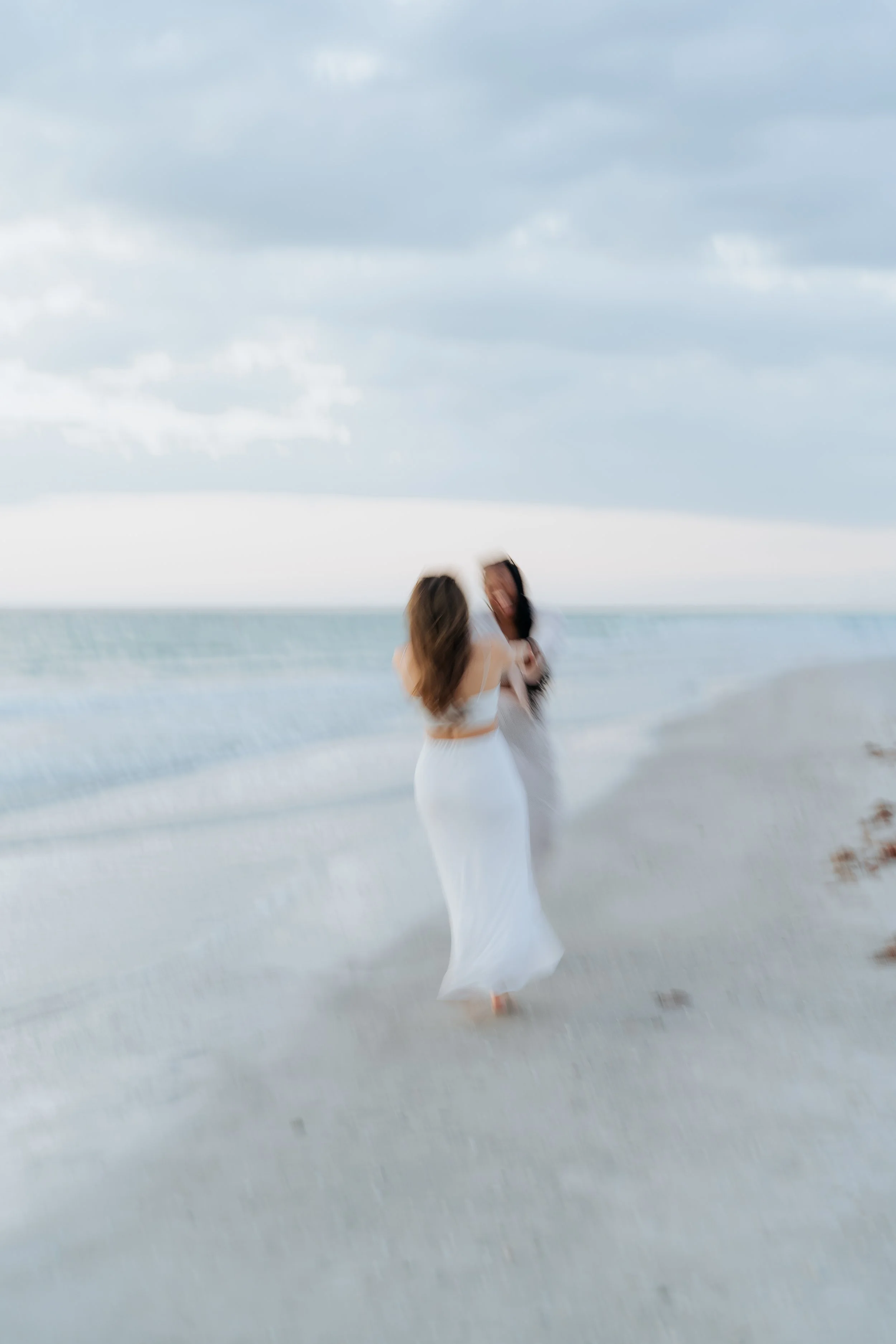 Two women in white dresses hugging on a beach with cloudy sky