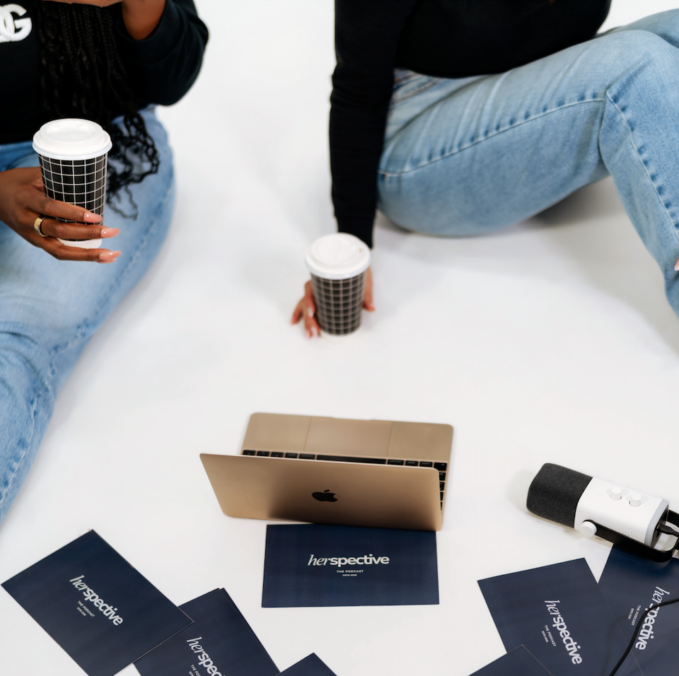 Two people sitting on a white surface with coffee cups, a laptop, a microphone, and several 'herpective' cards scattered around.
