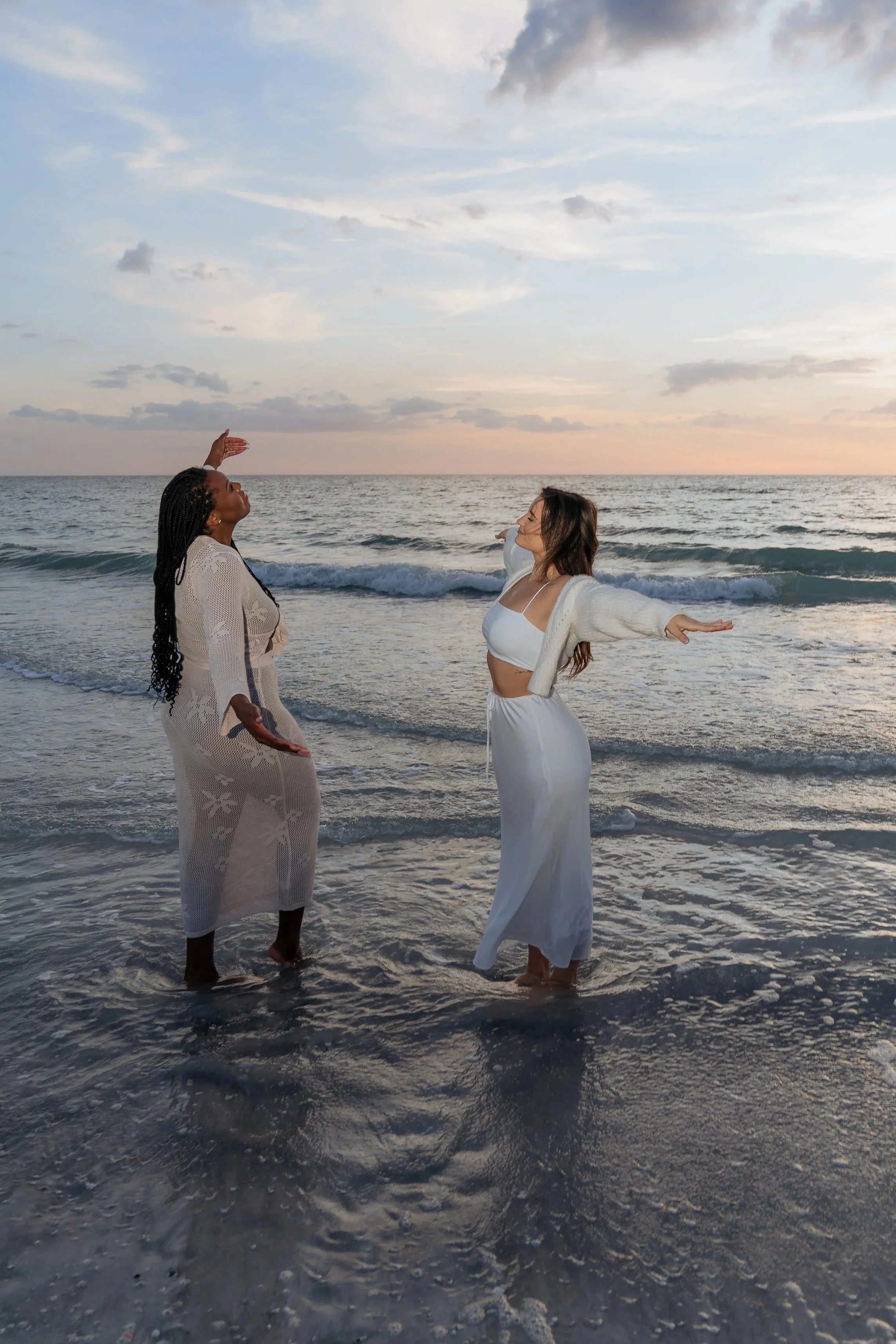Two women standing in shallow ocean water at sunset, with their arms outstretched, facing each other and the sky, wearing light-colored clothing, with clouds in the sky.