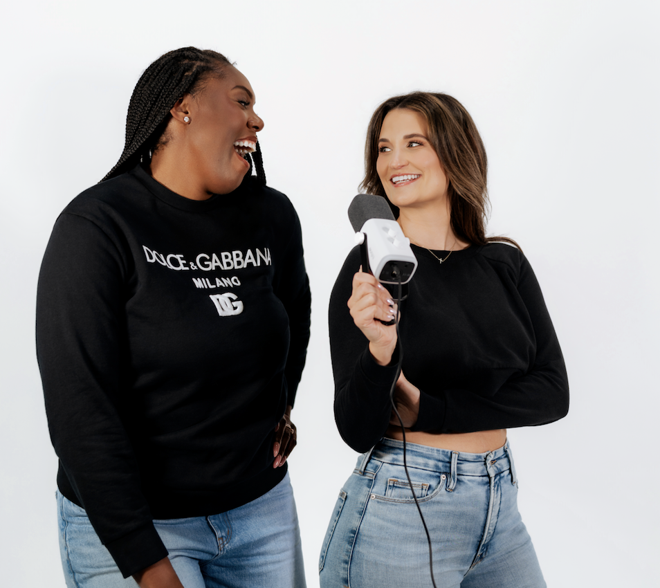 Two women laughing and talking during an interview, one holding a microphone, against a plain white background.