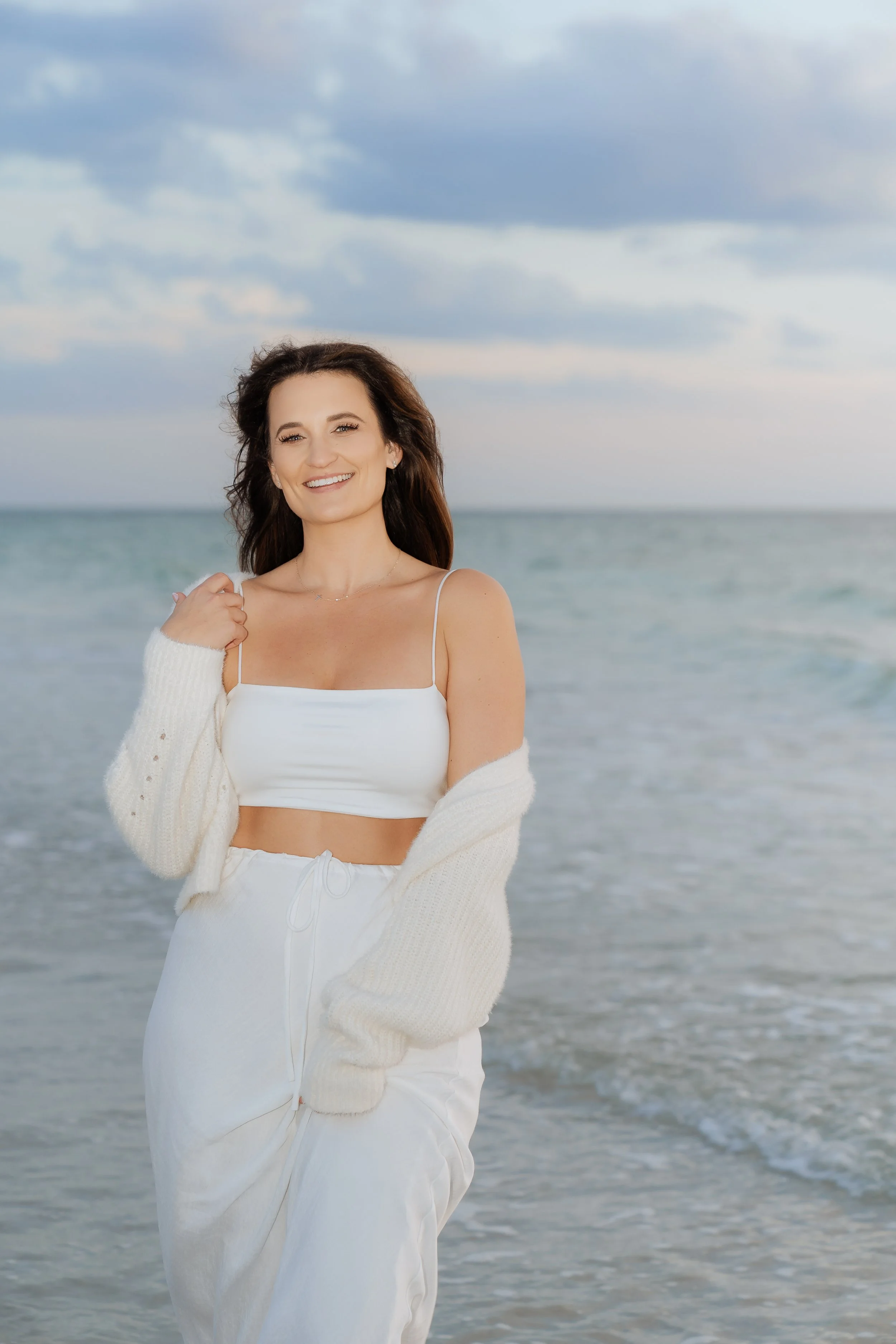 A woman with long brown hair and a white crop top is standing in front of the ocean with waves and a cloudy sky in the background. She is smiling and holding her sweater strap with one hand.