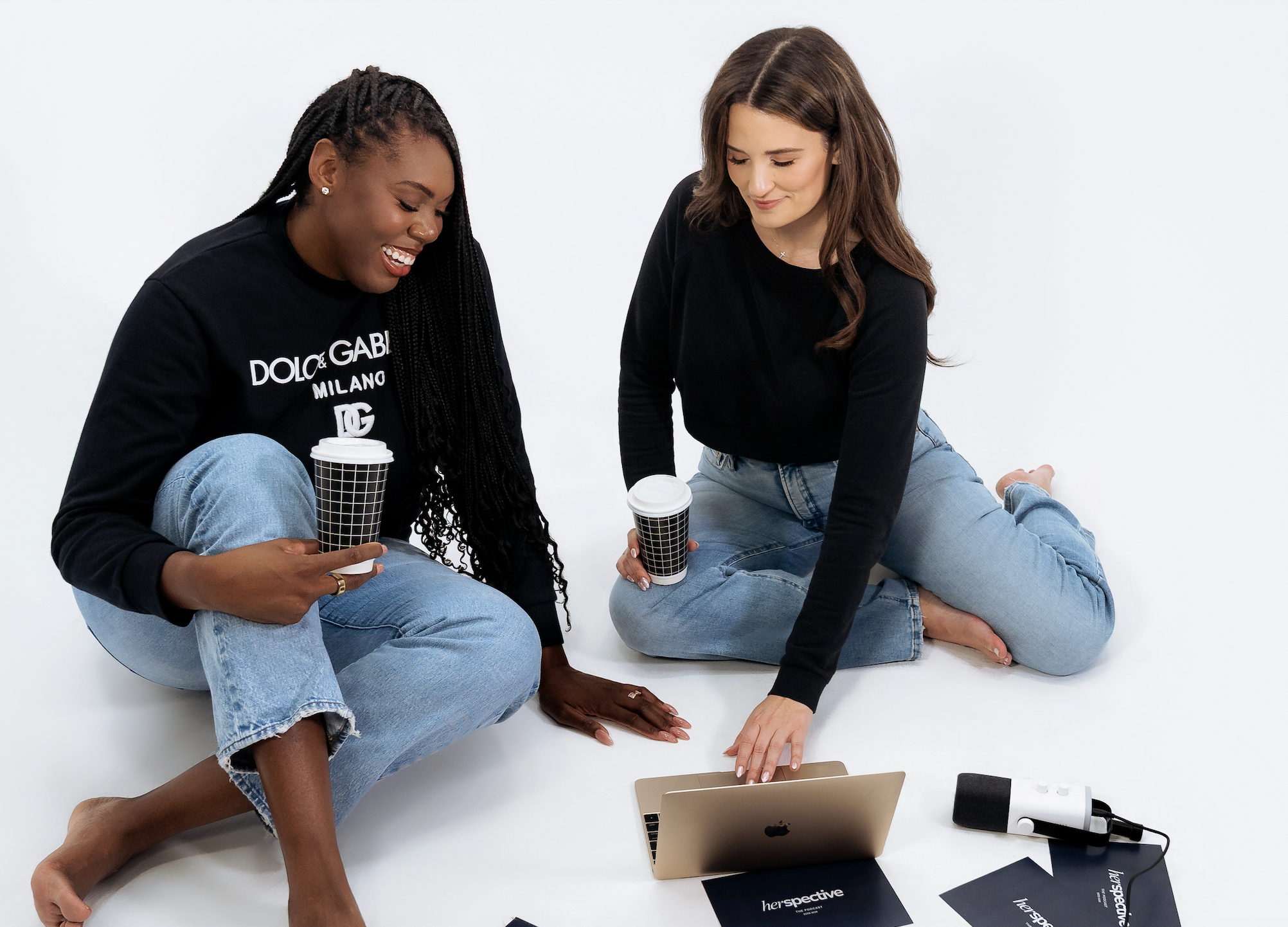 Two women sitting on the floor with a laptop, magazines, and a microphone, engaged in a discussion or presentation.