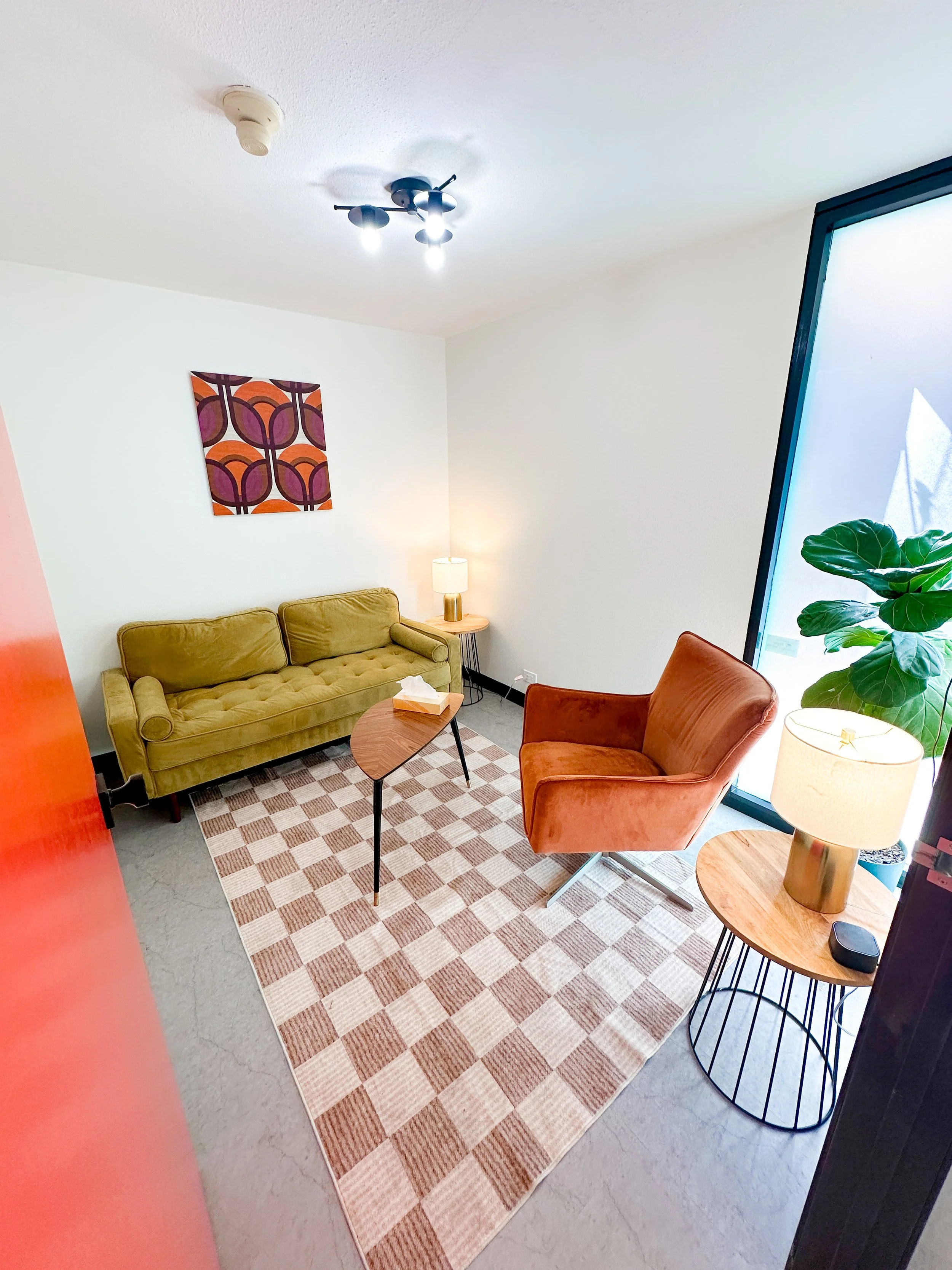 A cozy living room with a beige tufted sofa, an orange armchair, and two round wooden side tables with lamps. There is a colorful geometric painting on the white wall, a checkered area rug, a large green plant next to a tilted window, and ceiling-mounted lights.