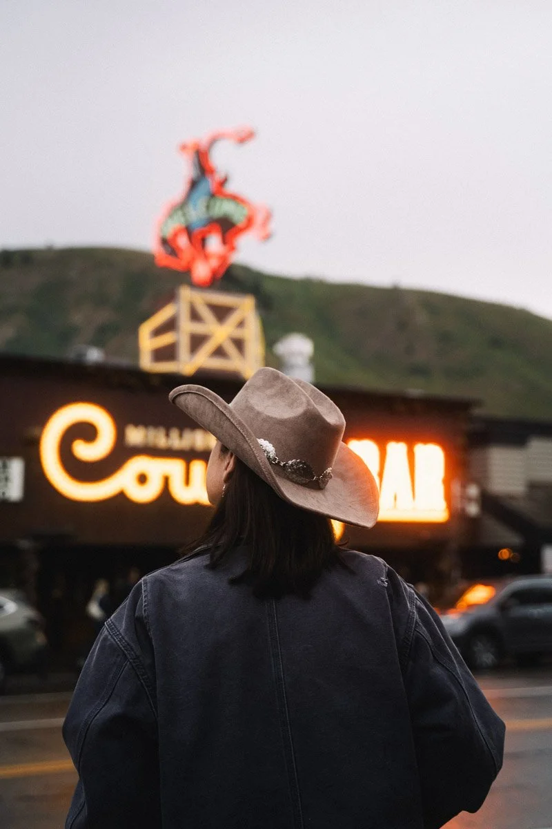 A woman with black hair wearing a brown cowboy hat and a denim jacket, facing away and looking at a neon-lit sign for a restaurant or bar, with a hill and a neon sign of a dinosaur in the background.