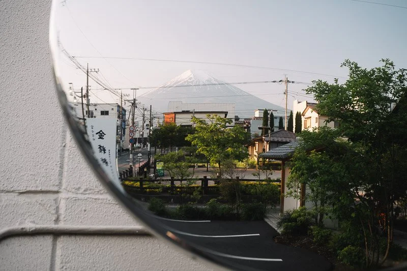 A view through a circular mirror showing a street scene with trees, buildings, and power lines, with Mount Fuji in the background.