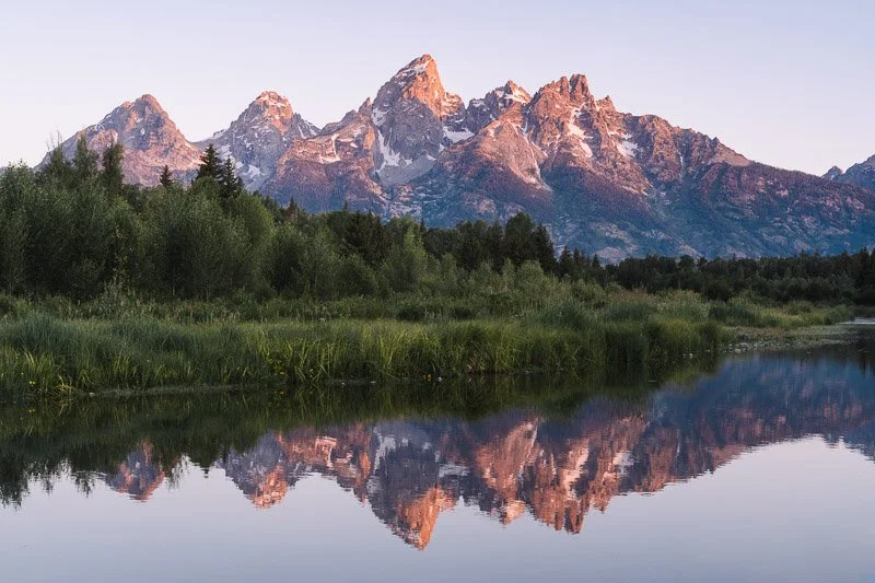 Snow-capped mountains reflecting in a calm river with green trees and grass in the foreground.