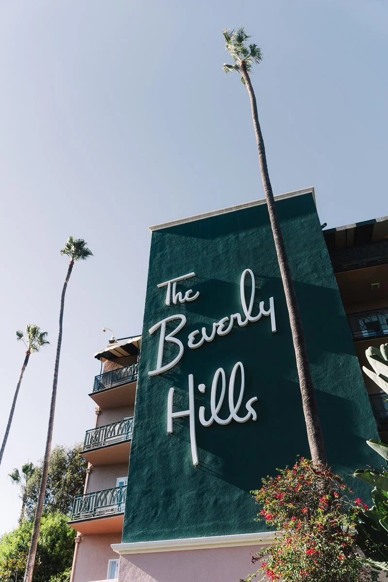 A large sign reading 'The Beverly Hills' on a green wall outside a building, with tall palm trees and a clear blue sky in the background.