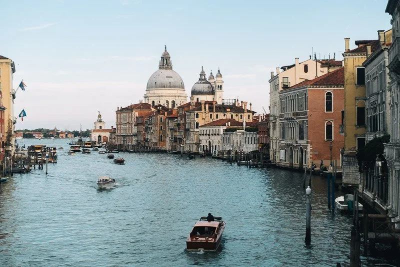The Grand Canal in Venice, Italy, with historic buildings along the water and boats, including a vaporetto, with the Basilica di Santa Maria della Salute in the background under a partly cloudy sky.
