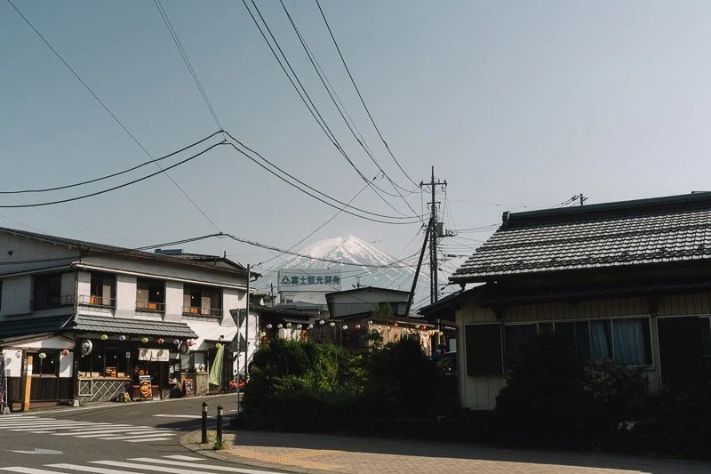 A street scene with buildings and power lines, Mount Fuji in the background, and a cloudy sky.