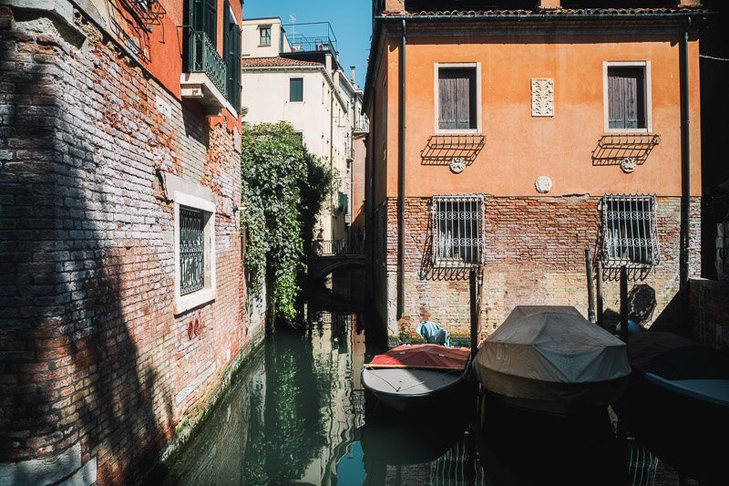 Venetian canal with three moored boats, flanked by old brick and stucco buildings with barred windows.