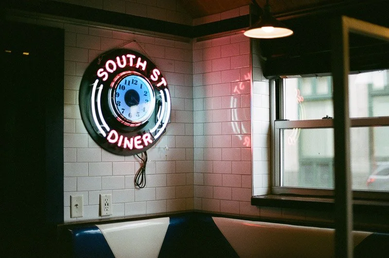 Neon sign reading 'South St. Diner' with a clock, inside a diner with a window and booth seating.