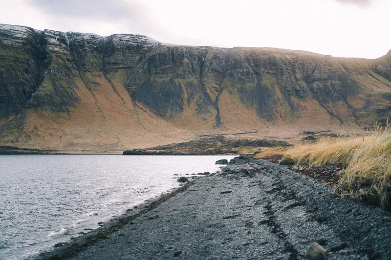 A rugged dirt road runs along a body of water with mountains in the background under a cloudy sky.