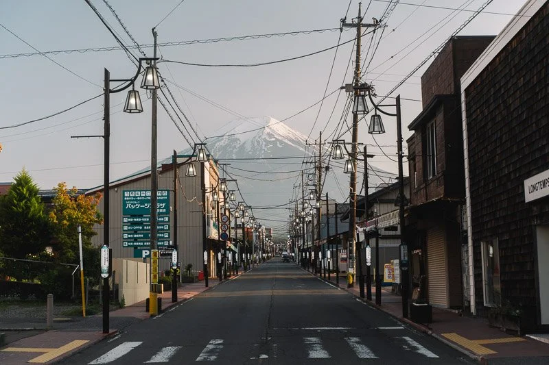 Empty street in a small town with buildings on each side, lampposts, and power lines, with Mount Fuji in the background.