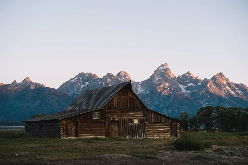 A rustic wooden barn with a sloped roof in front of a mountain range with snow-capped peaks.