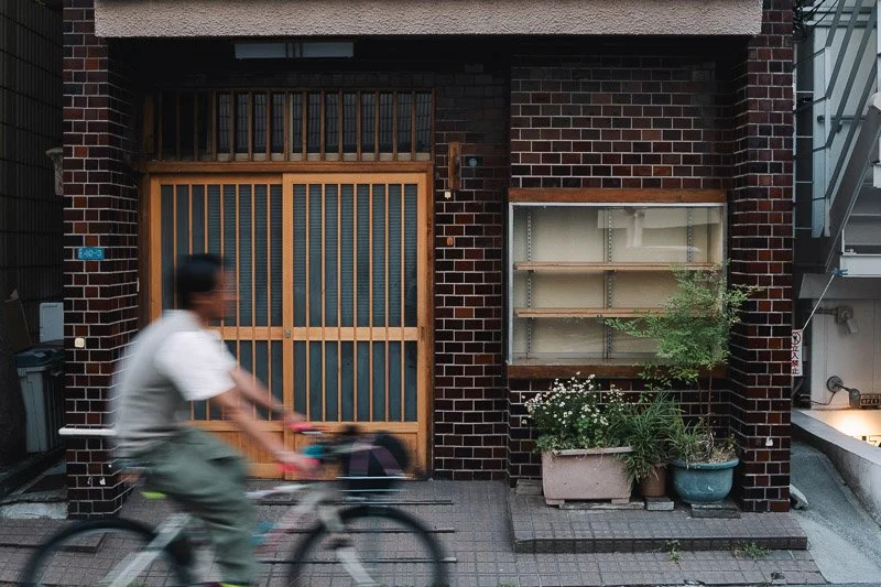 A person riding a bicycle in front of a dark brick building with a wooden sliding door and empty shelves surrounded by potted plants.