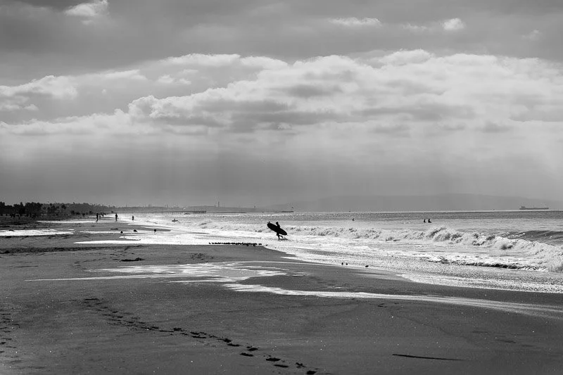 Black and white photo of a beach with people, waves, and a person carrying a surfboard walking along the shoreline.