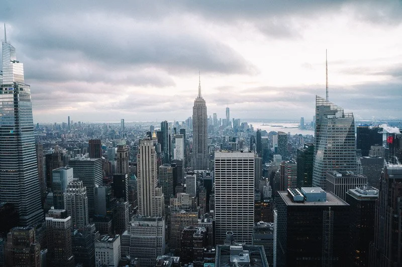 A panoramic view of New York City skyline featuring the Empire State Building in the center surrounded by tall skyscrapers under cloudy sky.