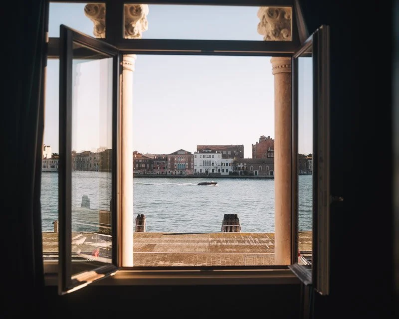 Open window looking out onto a waterway with buildings across the water and a boat passing by.