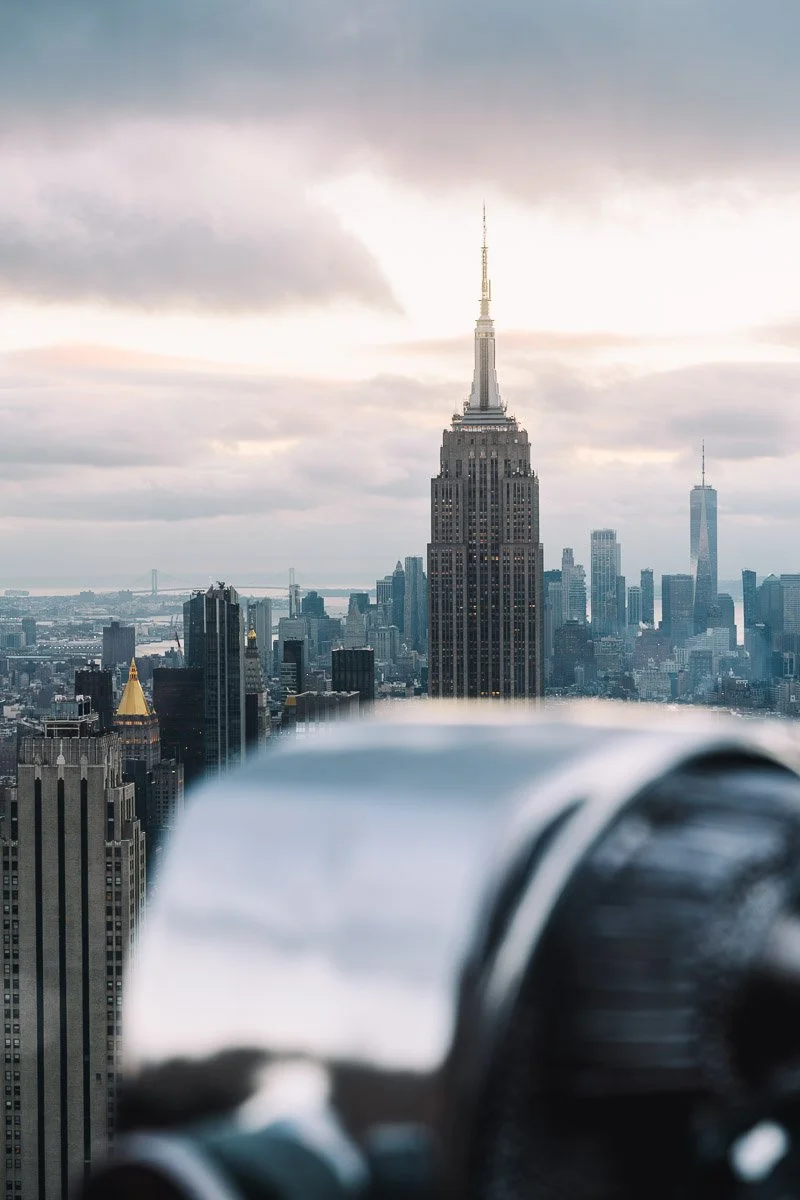 View of the New York City skyline featuring the Empire State Building in the foreground, with One World Trade Center in the distance, under a cloudy sky.