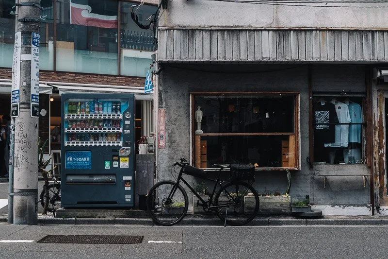 A bicycle parked next to a vending machine and a gray building with a shop window showing clothing inside.