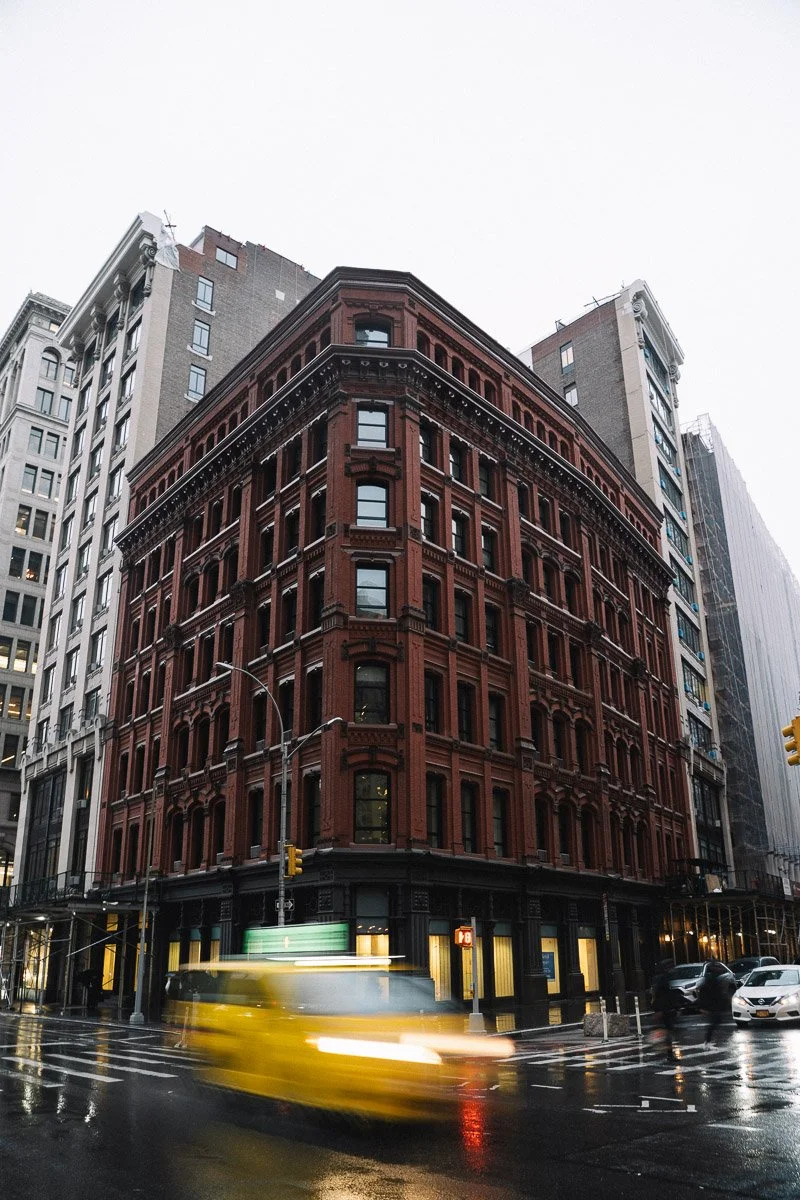 A corner view of a historic red brick building with detailed architecture in an urban setting, with other tall buildings surrounding it, on a rainy day with wet streets and a yellow taxi passing by.