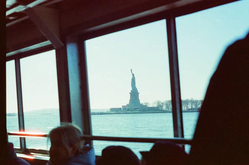 View of the Statue of Liberty through a boat window with passengers in the foreground.
