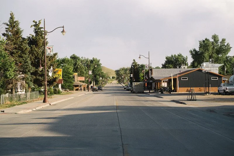 A small-town street scene with a wide road, trees, a few buildings, and parked cars, under a partly cloudy sky.