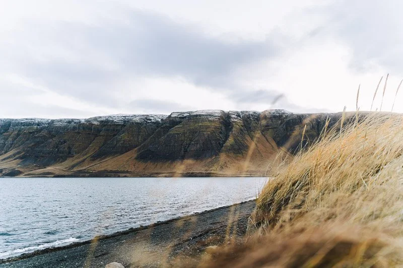 A shoreline with a lake or sea, rocky beach, grassy hillside, and mountains in the background under an overcast sky.
