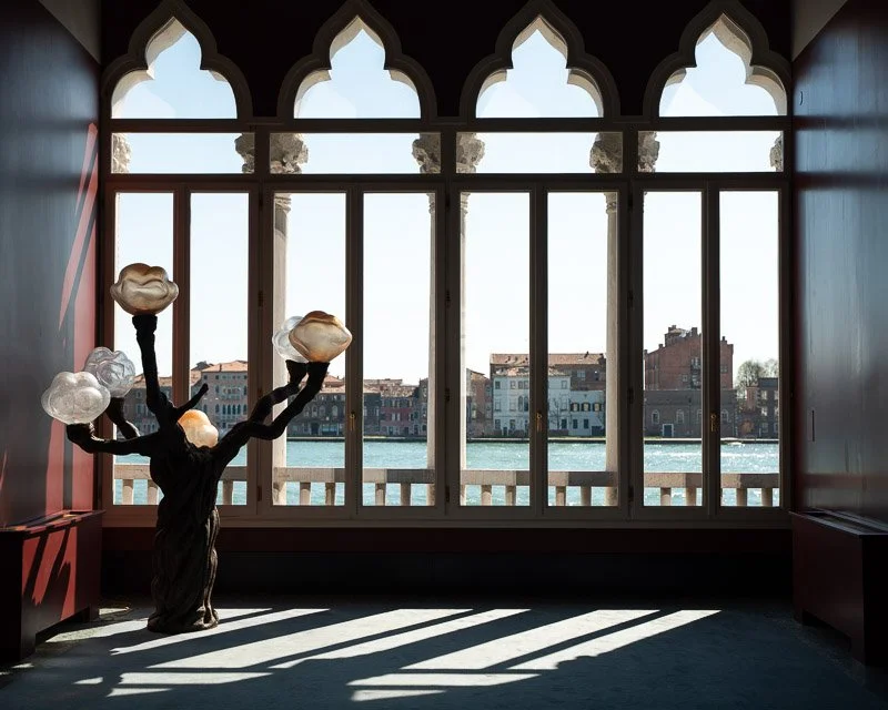 Interior of a room with large arched window overlooking a canal and buildings, a black sculpture of a hand holding four light fixtures shaped like clouds, and sunlight casting shadows on the floor.