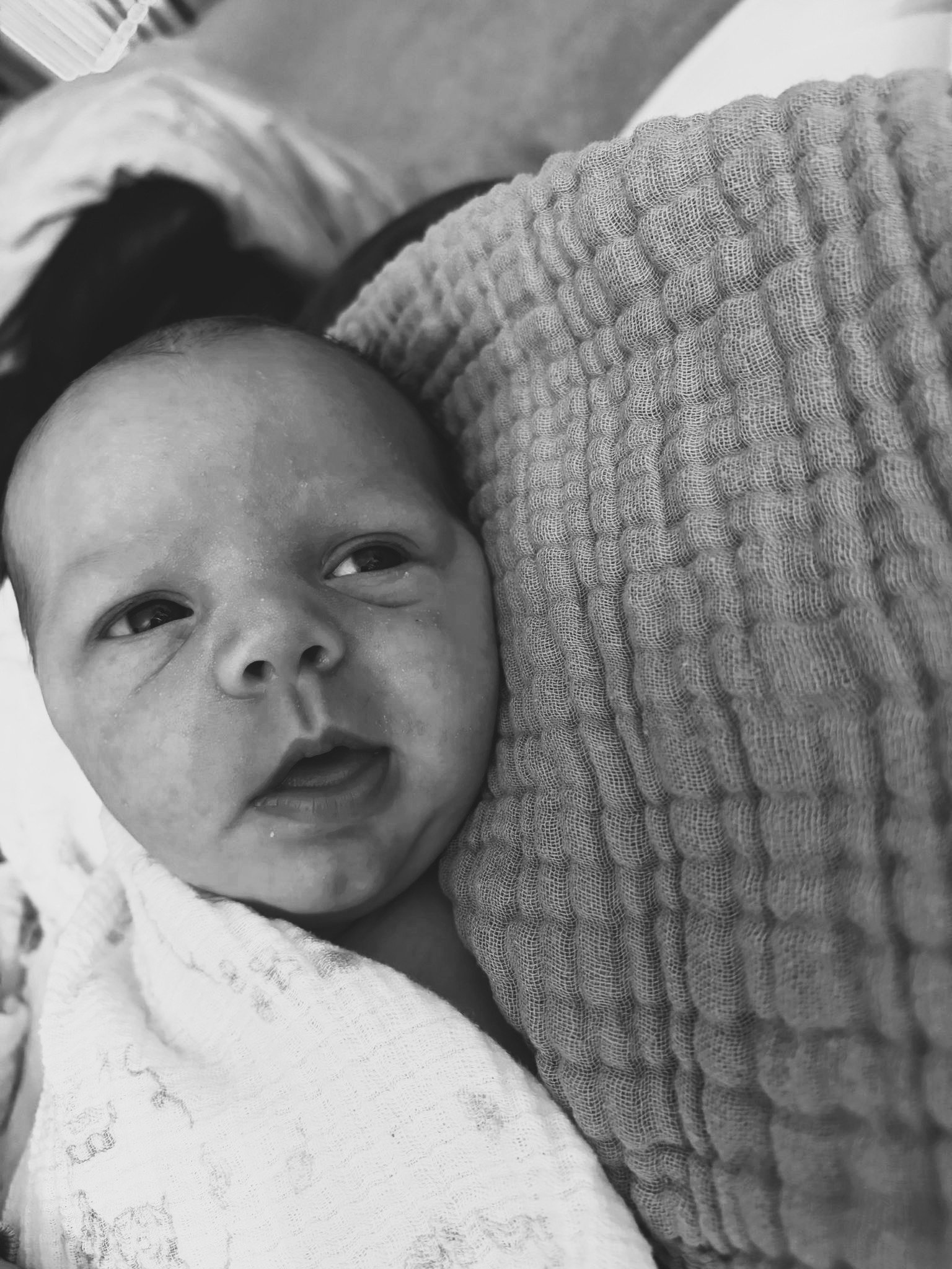 Close-up of a baby's face. The baby is wrapped in a light-colored blanket and appears to be lying down.