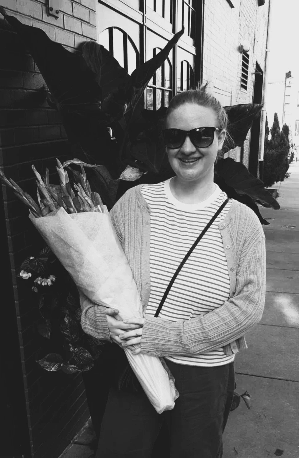 A woman smiling, wearing sunglasses, holding a bouquet of flowers, standing on a sidewalk next to a brick wall with plants and window decorations.