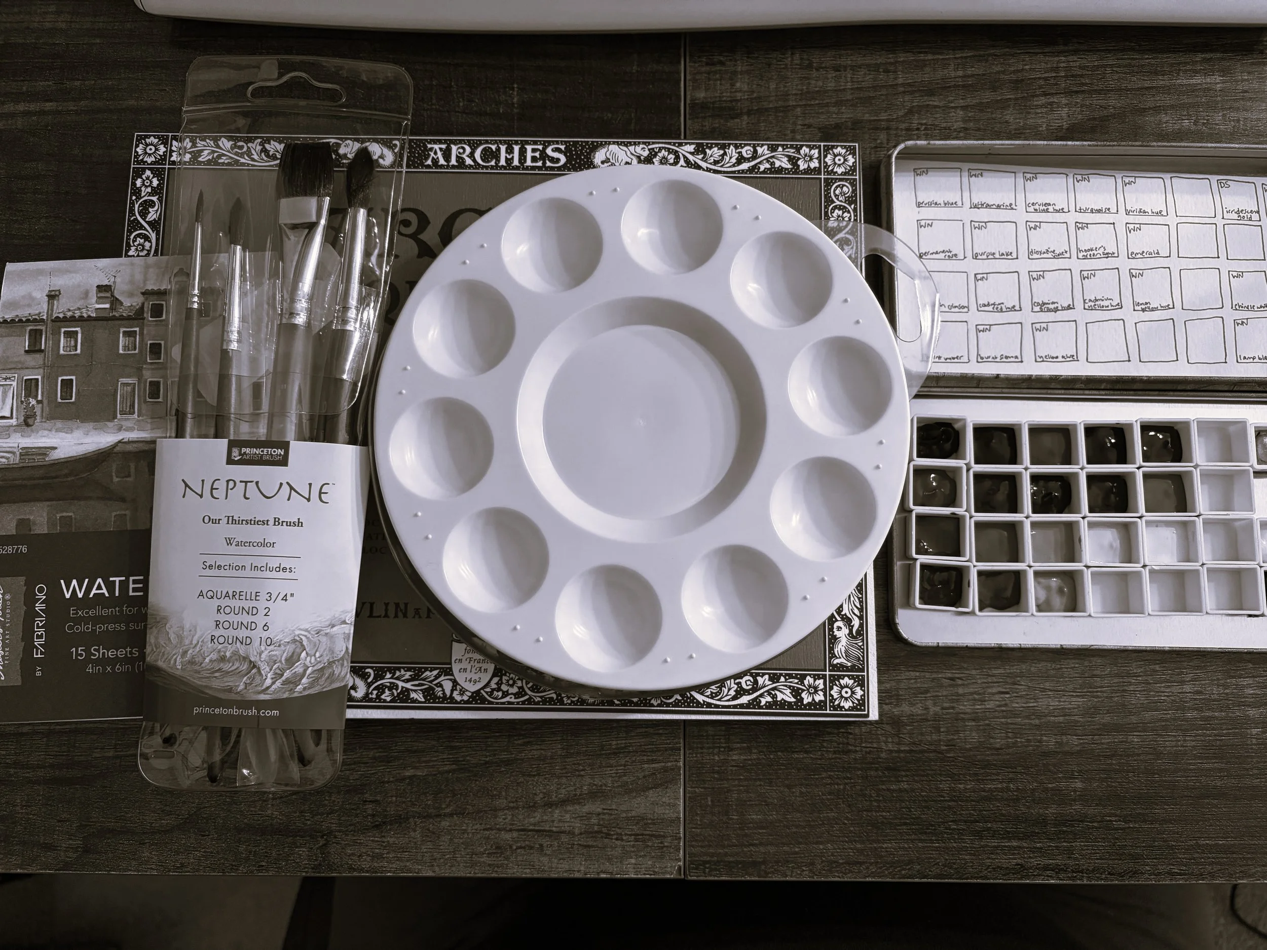 Art supplies including a white watercolor palette, a box of watercolor paints, a pack of Princeton artist brushes, and a watercolor paper pad on a wooden surface.